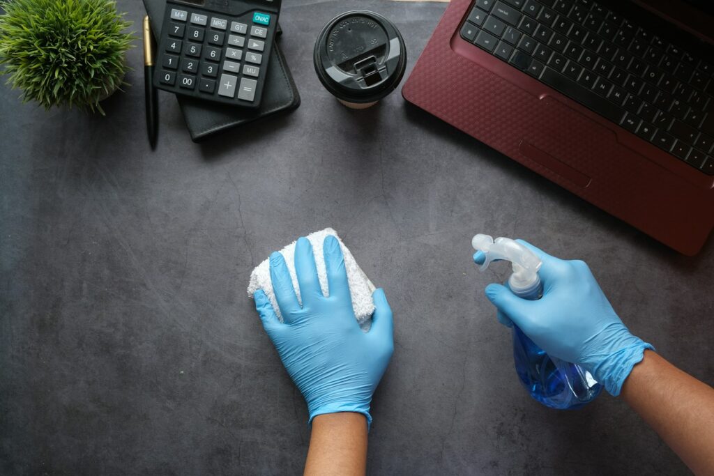 Gloved hands wiping down a dark desk with a spray bottle and cloth during cleaning